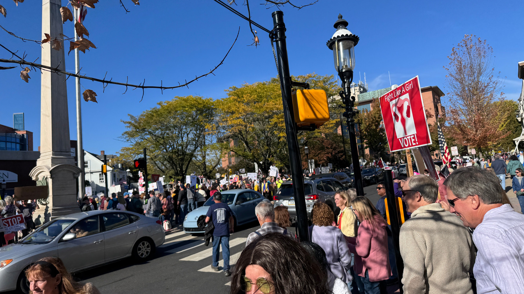 A large group of people is gathered at a busy street intersection, holding various signs and participating in a public demonstration.