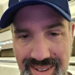 Man wearing a blue cap with the logo 'GM Goldstein Media', standing indoors in front of a structural background.