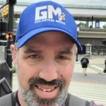 A man wearing a blue cap with 'Goldstein Media' logo taking a selfie at a crosswalk.