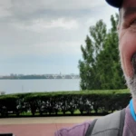 A man with a beard is smiling in front of a river with a bridge in the background.
