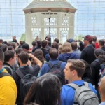 A large group of people gathered indoors, facing a speaker in front of an ornate structure at a conference venue.