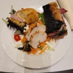 A plate of food featuring a sandwich, roasted meat with blackened crust, and salad.