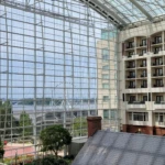 An atrium with a large glass wall overlooking a river and a bridge, with a hotel interior and small building visible inside.